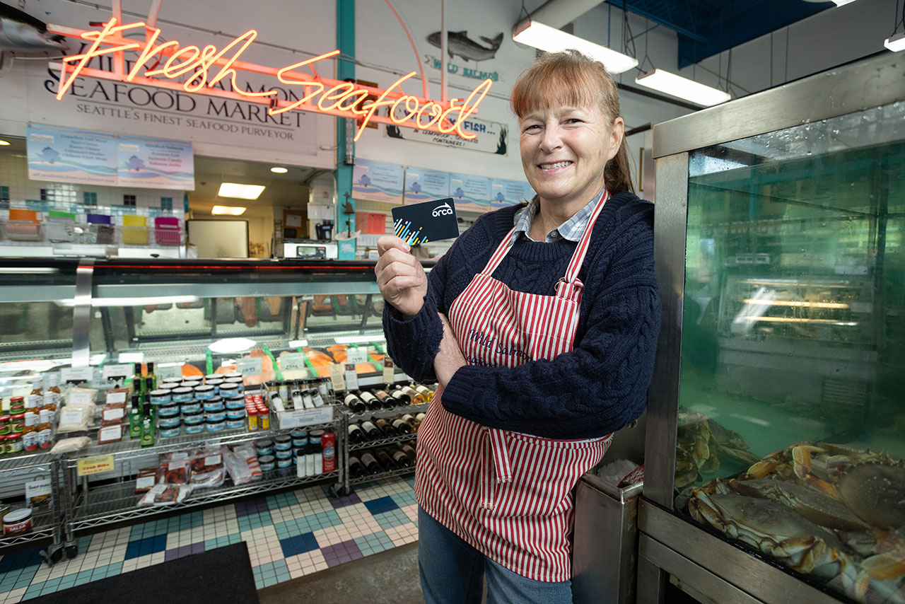 A person stands at a prep station in a pizza kitchen and holds up an ORCA card