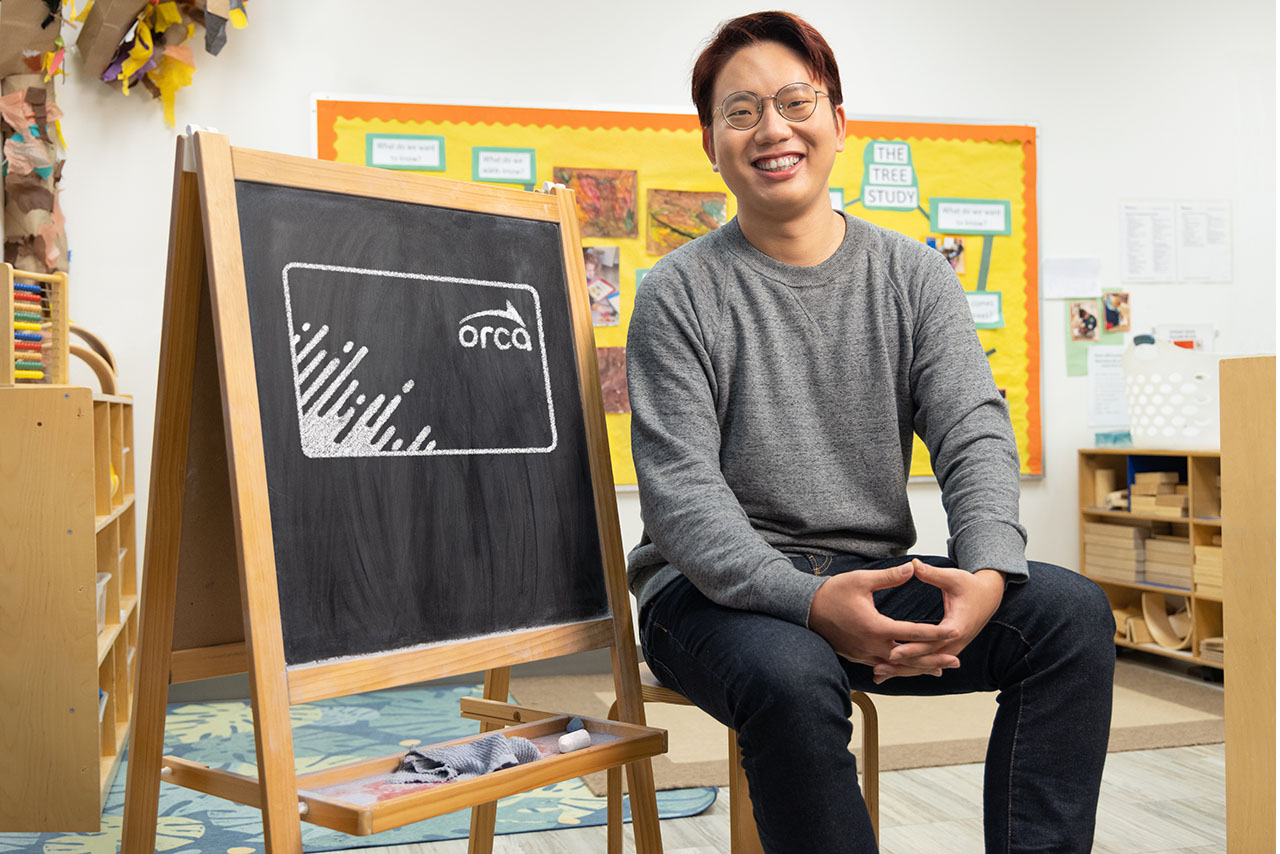 A person siting in a early childhoold education classroom. There is a blackboard with an ORCA card drawn on it in white chalk.