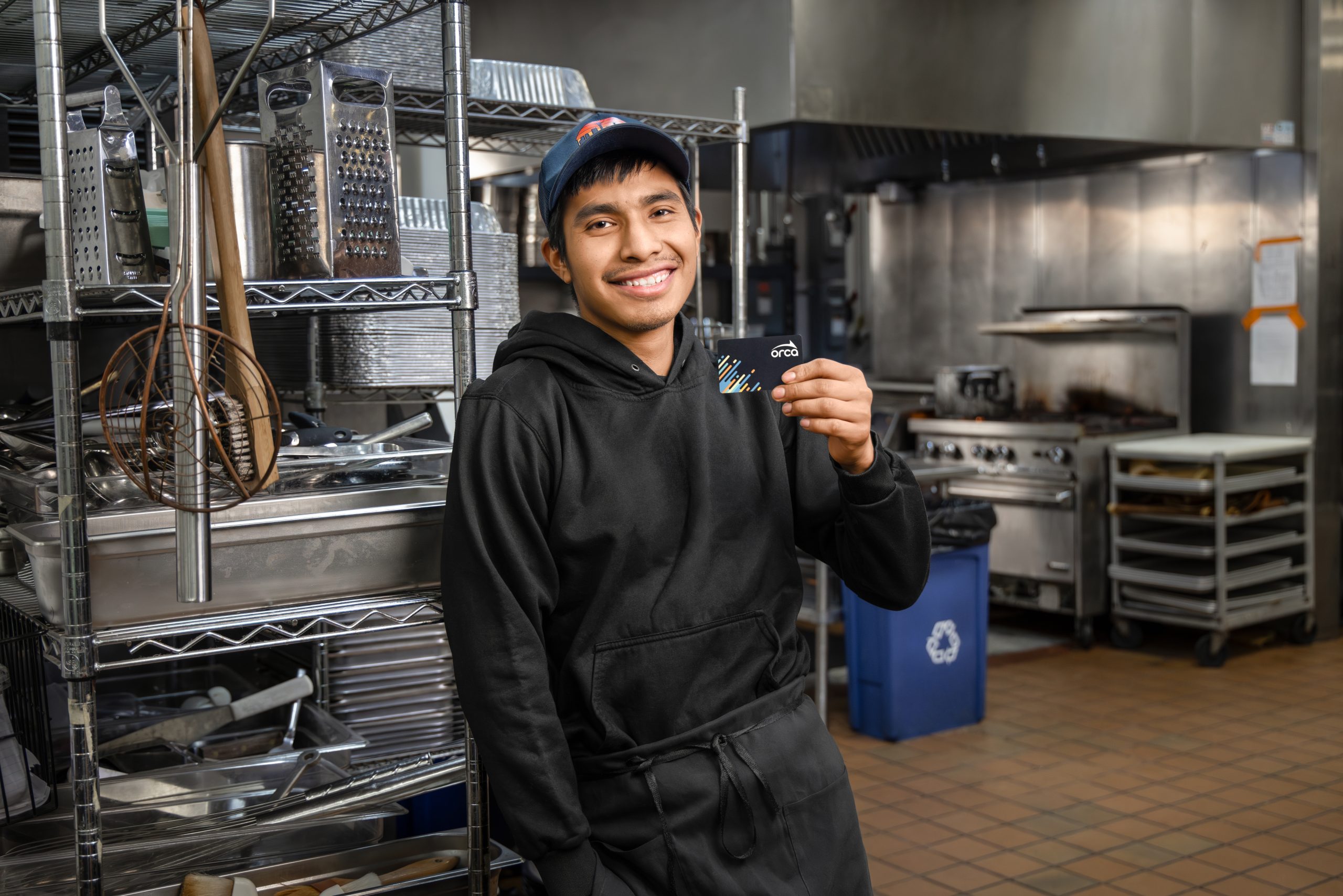 A restaurant worker standing in a commercial kitchen holds an ORCA transit card near metal shelving and cooking equipment.