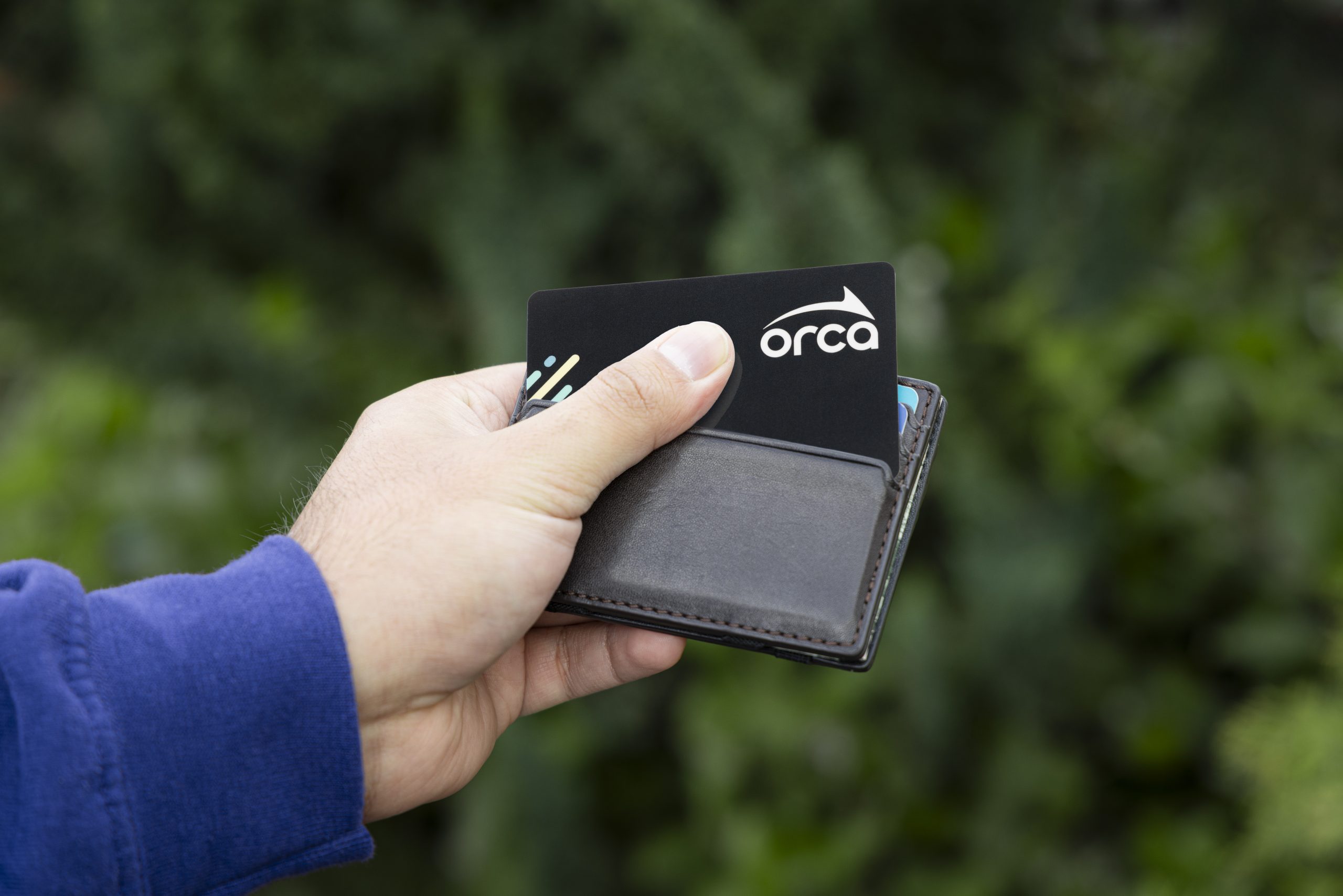 A hand holding a small wallet with a black “orca” card partially pulled out, set against a blurred green outdoor background.