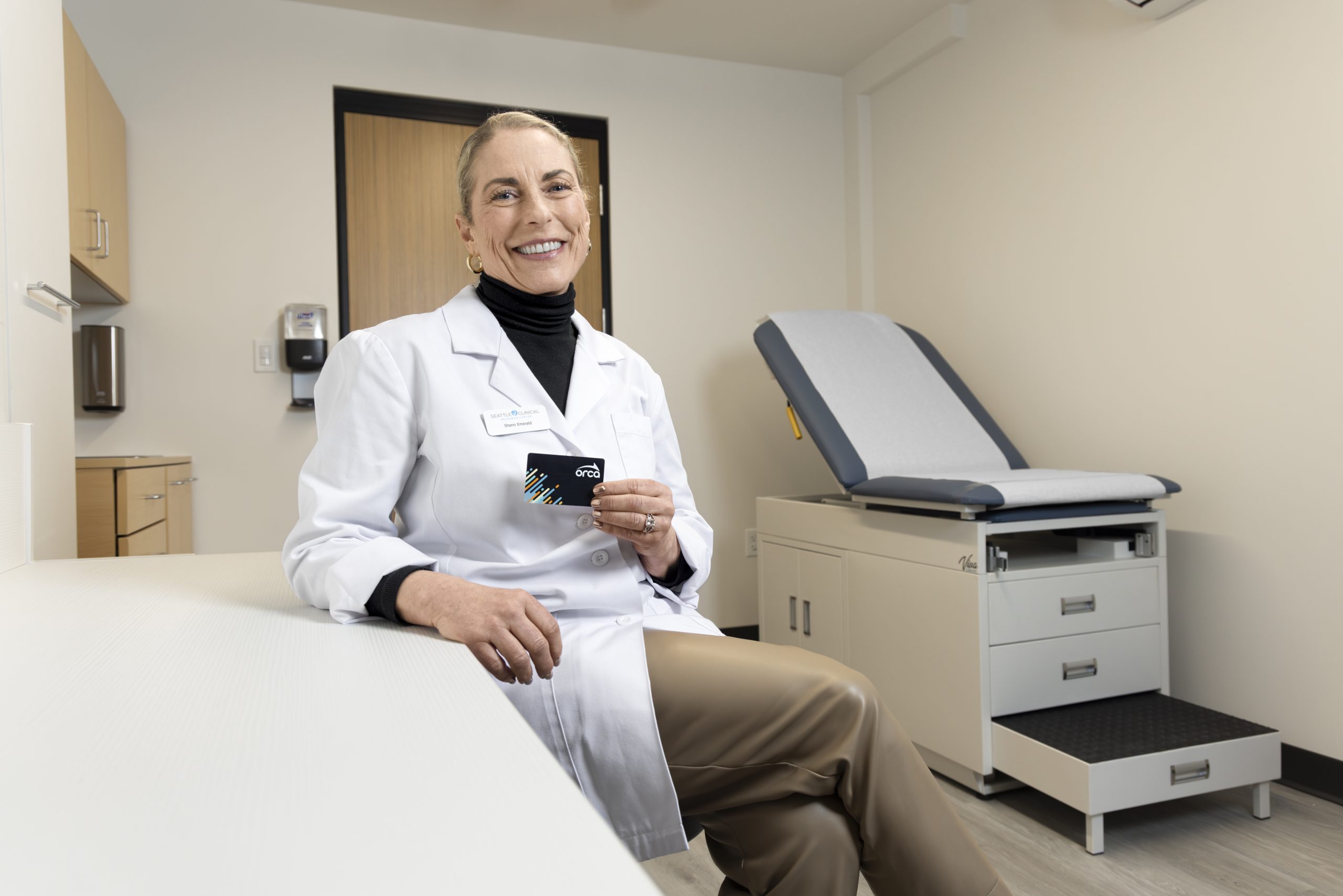 Person wearing a white lab coat seated on an exam table in a medical exam room, holding a small card, with an exam chair and cabinets in the background.
